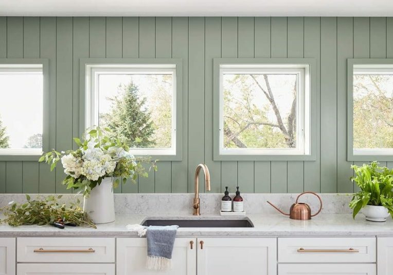 Modern kitchen with sage green vertical shiplap walls, white cabinetry with copper hardware, a white quartz countertop, and four white sliding windows over a sink
