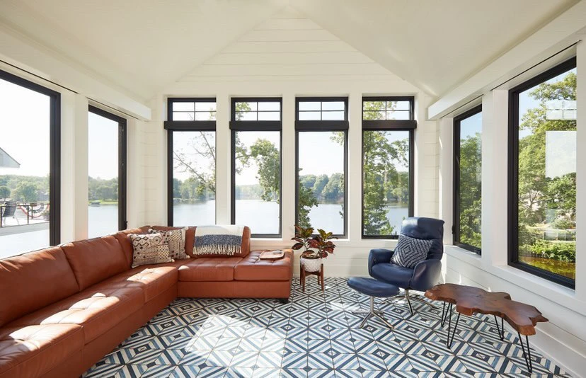 Modern sunroom with black-frame windows overlooking a lake featuring a tan leather sectional sofa and patterned tile flooring