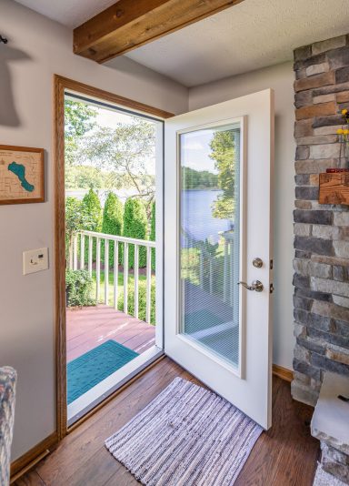 Interior view of an open ProVia white fiberglass entry door with a full glass lite, overlooking a wooden deck and a scenic lake surrounded by trees